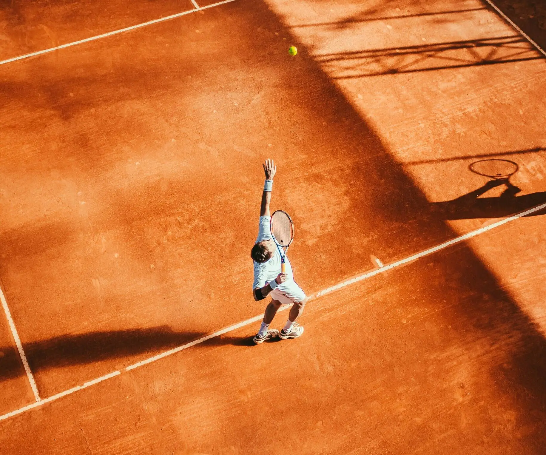 Aerial view of a tennis player serving on a clay court, highlighting a key skill developed in Evolve's science based tennis training and tennis high performance camp programs for effective learning tennis.